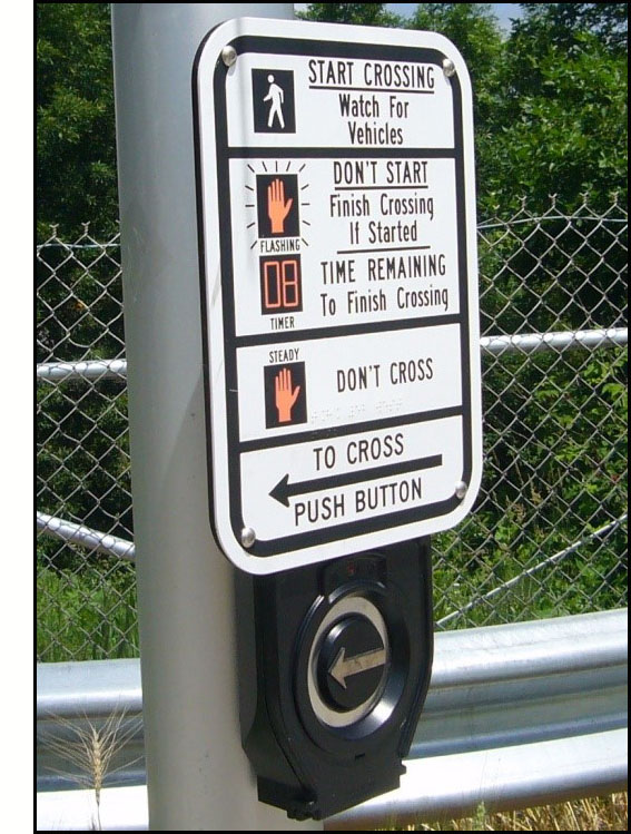 Photo shows a pedestrian pushbutton with a raised arrow on the button, pointing toward the crosswalk.