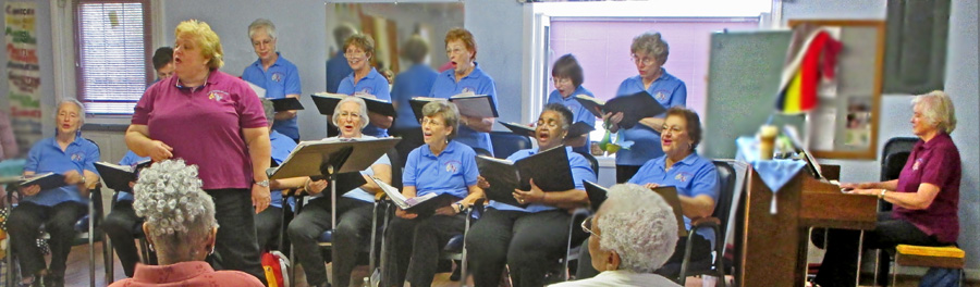 Photo shows Mary Kate behind her music stand, wearing a burgundy-colored top and black pants.  She is facing the audience and standing in front of 13 singers wearing matching blue tops and black pants, holding their music and singing -- 7 of them are sitting in a row and the rest are standing behind them.  At the end of the singers we can see Suzann sitting at the piano and facing Mary Kate, she is wearing a burgundy top.