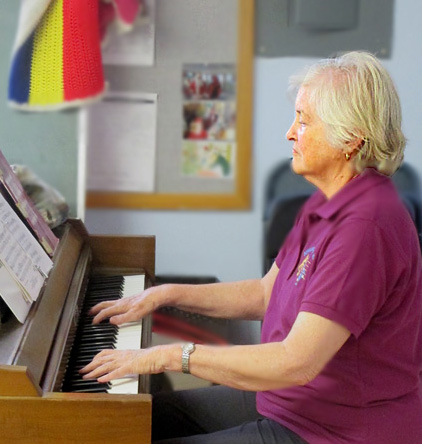 Photo shows Suzann wearing a burgundy-colored top and black pants and playing the piano.