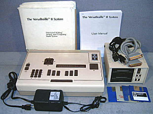 Photo shows the VersaBraille II with print and braille instruction books, cords, and a cube that looks like a disk drive, with slots on the front for disks.  The Versabraille II has the 6 braille keys and space bar, 3 rows of buttons that look like calculator numbers, a braille display of about 20 cells, and a few other buttons.