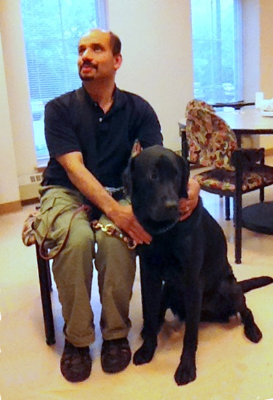 Bapin is sitting in a chair and smiling with his arm around a black lab dog who is sitting on the floor beside him.