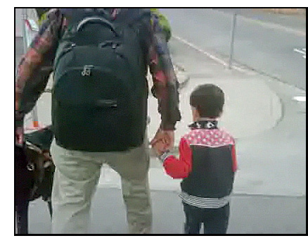 Photo shows the backs of Bapin and his son Navin as they walk across a street.  Bapin is holding his guide dog's leash with his left hand and Navin's hand with his right hand, and is wearing a very full backpack.