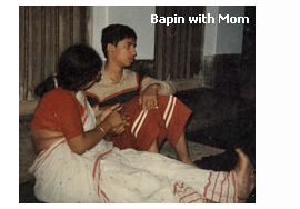 Photo shows Bapin, who appears to be a pre-teenager, and his mother sitting beside each other on a mat on the floor.  Bapin seems to have his back leaning against a wall that has curtained windows and is looking to his left. His legs are bent and his left hand is resting on his knees, his right hand is being held by his mother who is turned to lean and look toward Bapin.  She is wearing a burnt-orange short-sleeved shirt and a white sari trimmed with burnt orange, Bapin is wearing a button-down long-sleeved shirt with stripes, and loose burnt-orange pants with two white strips.