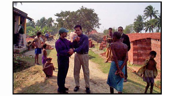Photo shows Bapin and a man both wearing long-sleeved buttoned shirts and dress pants standing on a narrow dirt road between stacks of bricks on one side and an open building on the left.  Bapin is also wearing a baseball cap, and smiling. His left hand on the man's right hand which is pointing toward another man who is approaching them, wearing no shirt and a blue sarang around his waist.  One child is walking beside them and looking toward the man with the sarang; several Indian children and adults are standing or sitting nearby, looking down the road behind Bapin at something we cannot see.  Two men are behind Bapin smiling and watching him interact, one has a shirt and a sarang around his waist, the other appears to have a shirt and pants.
