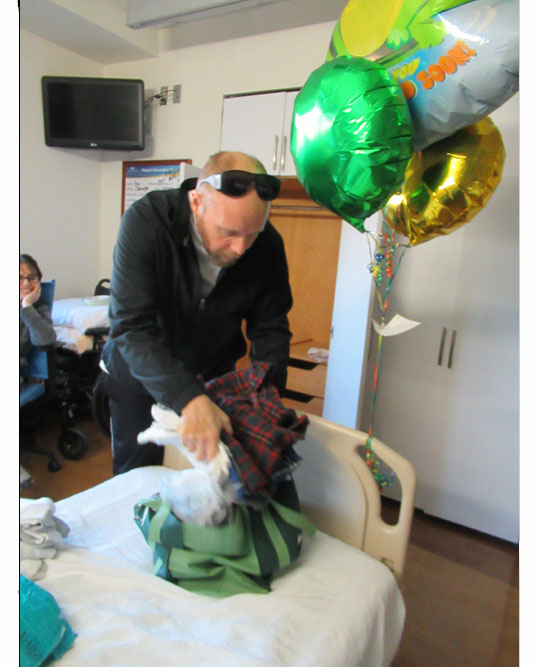 Two pictures show Paul standing at the foot of his bed with a cloth shopping bag, putting clothes into it.  Jomania is sitting on a couch behind him and watching, grinning in one of the pictures.  The large baloons are tied to the end of the bed.