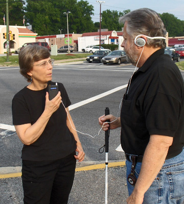 Dona is standing on a corner with a man who is holding a white cane.  The man is wearing ear phones connected by wire to a microphone which Dona is holding and talking into.