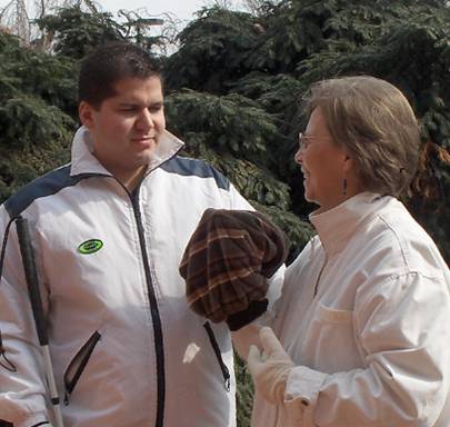 Photo shows Dona with a young man, both are wearing coats and smiling.  They each have one hand inserted in one end of the mobility muff, which is a fabric tube.
