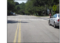 Photo shows a crosswalk on a two-lane street, with a car parked on the right (before the crosswalk).  A woman is standing at the edge of the crosswalk to our right, holding a cane up in the air.
