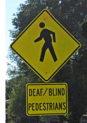 Photo shows a pole with two signs.  The top is shaped like a diamond and has a sillouette of a person walking.  Below that is a rectangular sign saying DEAF/BLIND PEDESTRIANS