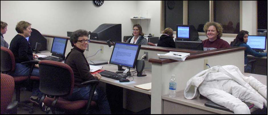 Photo shows 7 people sitting in front of 7 computers, 3 of them have turned to smile at the camera.