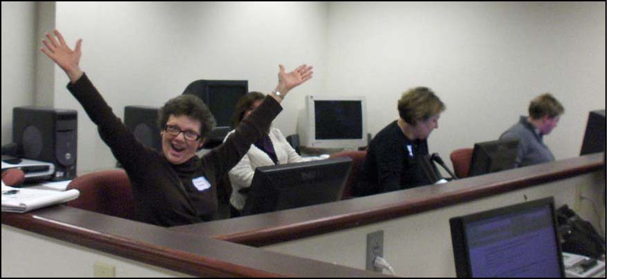 Photo shows one of the women at the computer, with a huge grin and both arms high up in the air.