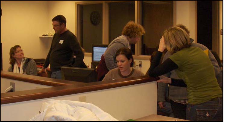One of the women who was laughing with the losers is now sitting in front of a computer, with two of the other women leaning over her shoulder.  Another woman sits at the next computer, and a man points to something on her screen.