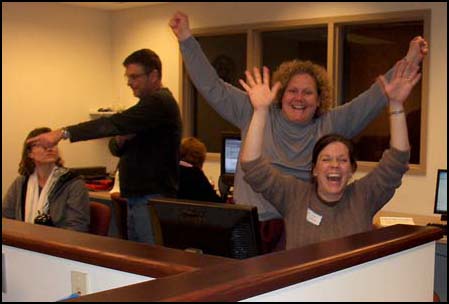 Photo shows two women in front of a computer, both laughing and triumphantly throwing their hands up.  Other women sit at other computers and a man points to something on one of their screens.