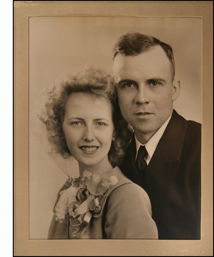 wedding photo shows a close-up of Bob and Jean, their heads close together, looking at the camera - Jean is smiling, Bob is smiling slightly. Jean's hair is softly curled around her face and a corsage of small orchids tied with ribbons is pinned to her suit.  Bob is wearing a very dark suit and tie.
