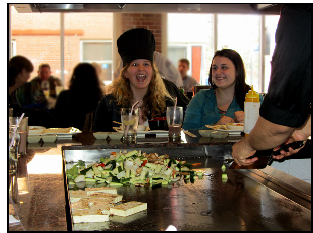 One of the students wears the chef's hat and opens her mouth while the chef tosses her a bite of food from the grill.