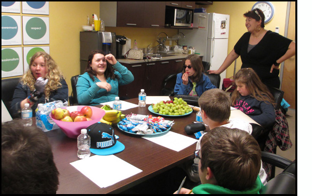 Photo shows students sitting around the table with snacks, eating and talking.  Dominique is standing and listening to one of the students.