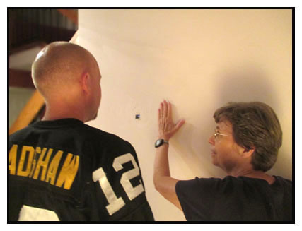 Two photos show Dona with a man whose head is shaved and who is wearing a Steelers football jersey, standing about two feet from a plain, light-colored wall.  The man is looking at a black spot on the wall, and Dona's hand is to the right of that spot, moving closer to it.