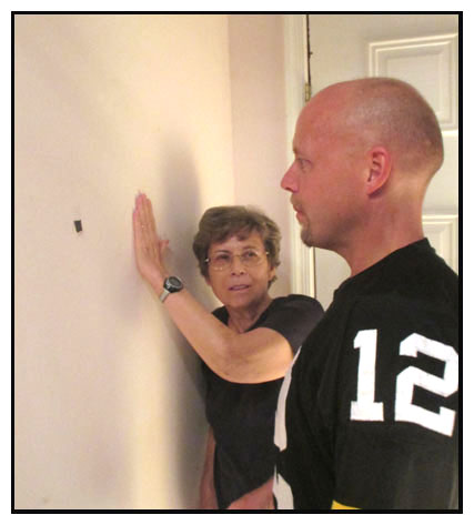 Two photos show Dona with a man wearing a Steelers jersey, standing about two feet from a plain, light-colored wall.  The man is looking at a black spot on the wall, and Dona's hand is to the right of that spot, moving closer to it.