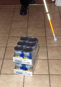series of photos shows a person walking toward a stack of cans (one six-pack on top of another).  The cane starts behind the pile to the person's left, and as it moves to the right it barely misses the pile and goes around the right side of the pile.  when the cane reaches to the left again, it is beyond the pile and misses it altogether.  The person continues walking into the pile.