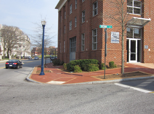 Photo shows a corner of an intersection of two 2-lane streets made of concrete; the sidewalk and the 3-story building at the corner are made of red brick.  The bricks slope to form a curbramp at the apex of the corner, with white cement detectable warning surface at the bottom.  A row of orange-colored bricks draws a rectangle that is about 4 feet wide at the bottom of the ramp and extends about 8 feet into the sidewalk.