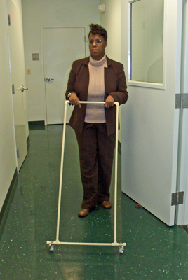 Three photos show the woman approaching the same door but holding an AMD instead of a cane.  The AMD is in the shape of a rectangle with wheels on the bottom, and she is pushing it like one would push a lawnmower.