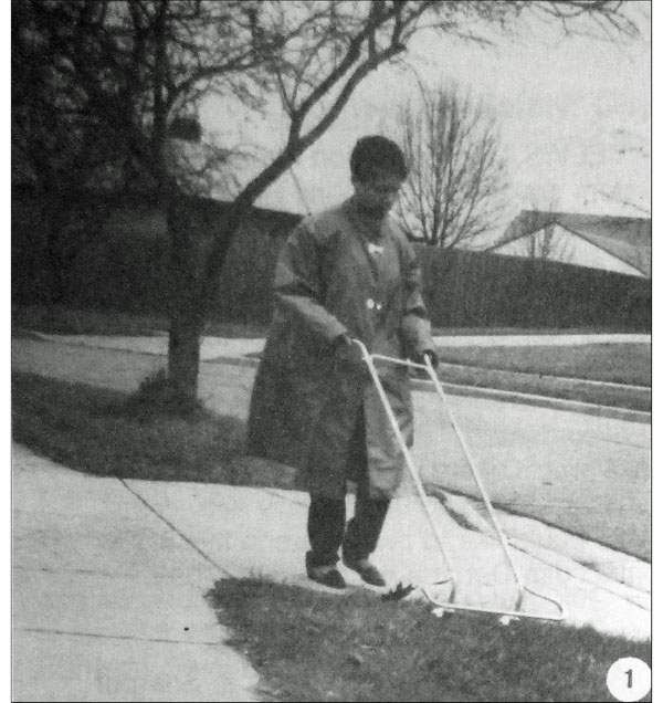 Photo shows woman wearing a light coat walking across a driveway.  She is pushing a rectangular AMD frame with wheels on the bottom, and it has reached the grass between the sidewalk to her right and the street to her left.
