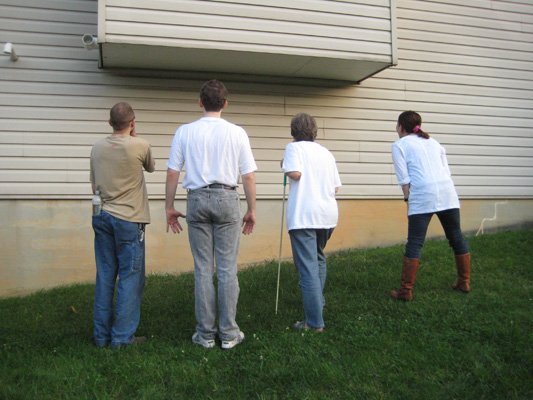 Photo shows everyone standing in the grass facing the side of a building about 3 feet away.  No one has a cane or AMD except Dona.  Mike and Stephan are standing straight, Mike is cupping his hands to his mouth.  Dona and Hiromi are leaning toward the building.
