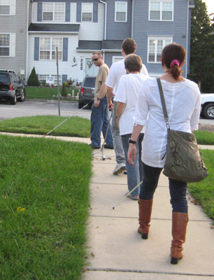 Photo shows Mike, Stephan, Dona and Hiromi walking in single file along a sidewalk.  Mike, at the head of the line, is using a cane and turning the corner, tilting his head toward the others.  Stephan is following close behind, pushing a wheeled AMD in front of him.  Dona is next and then Hiromi, they are both using a cane.