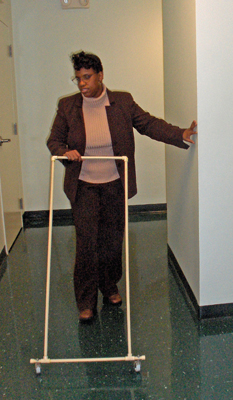 Photo shows the woman with her left hand trailing a wall approaching a corner, and pushing the AMD with her right hand.