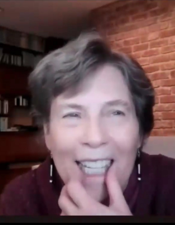 Dona Sauerburger is a white woman with short, dark blond hair, wearing earrings that look like white canes.  She is smiling and stroking her chin as she speaks.