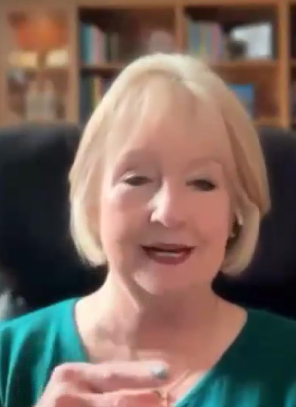 Laura Bozeman is a white woman with light blond hair cropped at chin length, wearing a green top, she is smiling and raising her hand while speaking.
