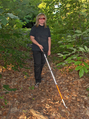 Two photos show a woman wearing black slacks and short-sleeved shirt walking through a green woods along a path of fallen leaves.  She is moving a white cane with a bundu basher tip along the ground in an arc from side to side in front of her. As it reaches the extent of the arc at each side, the tip is turned to face the center again.