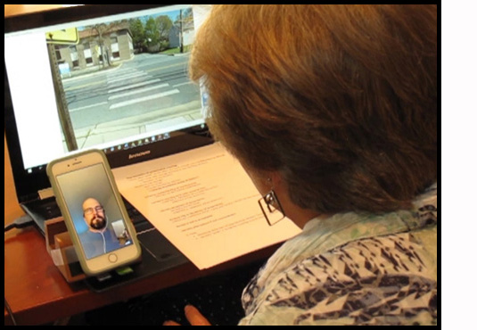 Picture shows Dona sitting at her computer, with a picture of a crosswalk on the screen and a checklist on the keyboard.  She is looking at Facetime on her smartphone, where she can see her student.