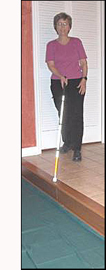 Dona has her right foot foward and the cane on her left, with the tip of the cane being at the edge of a drop-off that is diagonally (about a 45-degree angle) on her right.