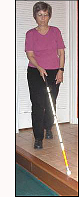 Dona has stepped forward so her her left foot is in front and the cane has moved to her right, and the tip of the cane is still at the edge of the drop-off that is diagonally (about a 45-degree angle) on her right.