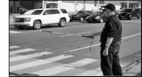 The figure is a photograph that shows a pedestrian wearing dark glasses and a baseball cap standing on the corner of an intersection at a zebra crossing. His long cane (which appears to be white in color with a red tip) is extended parallel to the ground from about his midsection.