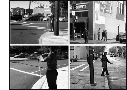 All 4 photos show a man wearing dark pants and shirt and a dark cap standing at different corners, facing one street with another street on his left.  Top left photo shows him facing a 7-lane suburban street -- with his right arm extended across his body, he holds a white cane with the grip near his left shoulder and tip at the curb in front of him.  Top right photo shows him in front of a large building with the cane extended about knee-high.  Bottom right photo shows him at the same corner stepping out into the street, with the cane extended about knee-high in front of him.  Bottom right photo shows a different man with cane held like the first man held it, and his left arm is extended to the left with the palm facing away from him.