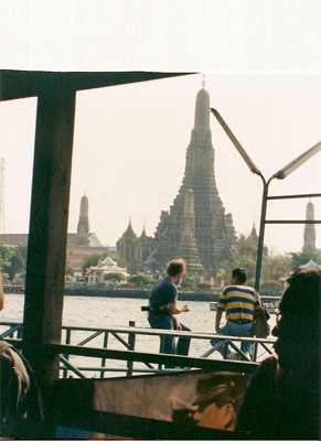 We are looking across the river to a bell-shaped temple ('wat) -- sitting on a railing at this side of the river are two men looking toward the river -- the one on the left is tall and has light hair (David).