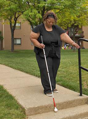 photo shows the woman's cane is over the edge of a stairway at the end of a sidewalk.  She is bending her head down to look at the edge and reaching her free hand to search for the railing.