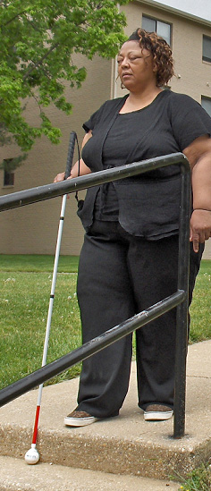 Woman stands with straight posture and eyes closed near the edge of the stairs, one foot forward, holding her cane at the edge.