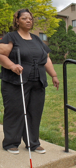 The woman approaches the stair, her cane has dropped over the edge and she is standing with good posture, looking forward.