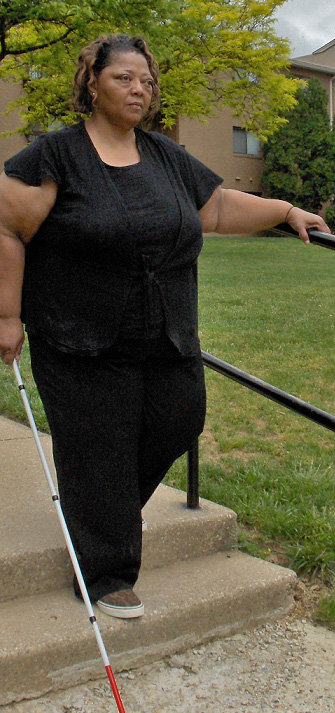 The woman has stepped down to the first step, her hand is on the rail and her other hand holds the cane at her side, extended to reach the landing.  SHe is looking forward, and standing straight in good posture.