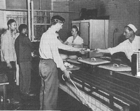 two blinded soldiers in a cafeteria line, with instructor Dee Corbett watching.  The first soldier holds a long white cane with a large crook in one hand and with the other he reaches over the counter for a plate that is being handed to him by a worker.