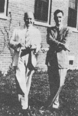 Russ and Warren stand beside each other in front of the side of a building, smiling and wearing a suit and tie.  Their legs are crossed, Russ's arms are crossed, and two fingers of each hand is crossed.