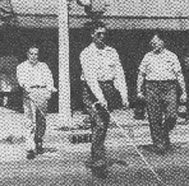 Two blinded soldiers cross a street, walking about 10 feet apart and canes each extending diagonally in front of them.  Instructor Eddie Meese walks beside them, watching.