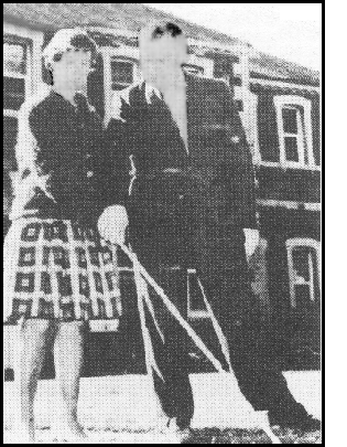 photo shows middle-aged Richard Hoover wearing a coat and tie, standing next to a young blind woman who is wearing a sweater and plaid skirt, she has a white cane extended in front of her on what appears to be a campus lawn, and he has his hand on hers