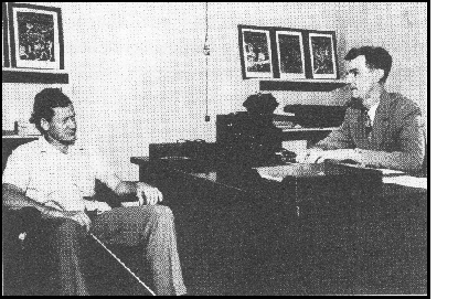 photo shows Russ Williams wearing a coat and tie and sitting behind a desk, reading a braille paper and speaking to a man who is sitting in a chair in front of the desk, wearing slacks and a button-down white shirt with no tie, holding a long white cane, and listening to Russ.