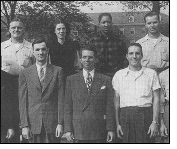 Photo shows one woman and 6 men and part of a 7th man posing outside under some trees with a large building in the distant background.  Russ and the man next to him are wearing a coat and tie, the other men are wearing button-down shirts with top button open and no tie (the shirts appear to be identical white shirts except for Dee's shirt, which is plaid).  The woman is wearing a dark dress.  Dee Corbett is African-American, all other people in the photo are Caucasian.  No one is holding any cane.