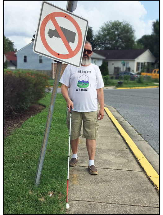 photo shows a man with a white cane walking on a residential sidewalk and approaching a sign that has been tilted into the sidewalk.  If he continues to walk straight, the cane will go under the sign and he will hit it with his head.