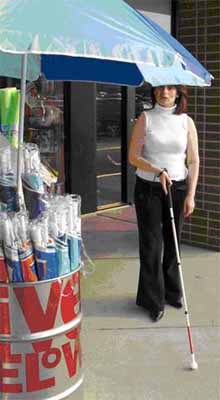 photo shows a woman using a white cane and walking along a sidewalk in front of a store.  A beach umbrella is opened and the woman is about to walk into it and bump her head.